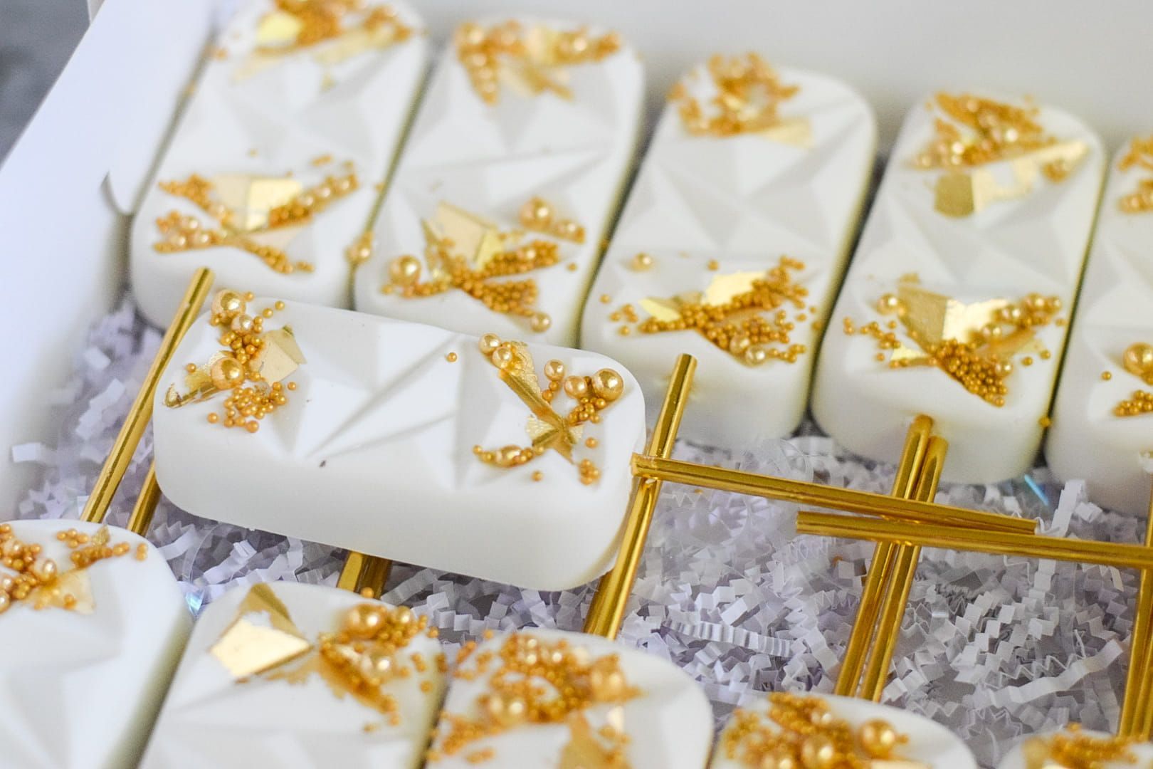 White rectangular cake pops with gold sprinkles and geometric designs in a box.