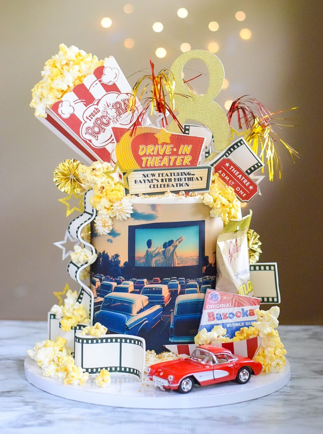 Drive-in movie themed cake with popcorn, a red car, and film reel decorations.