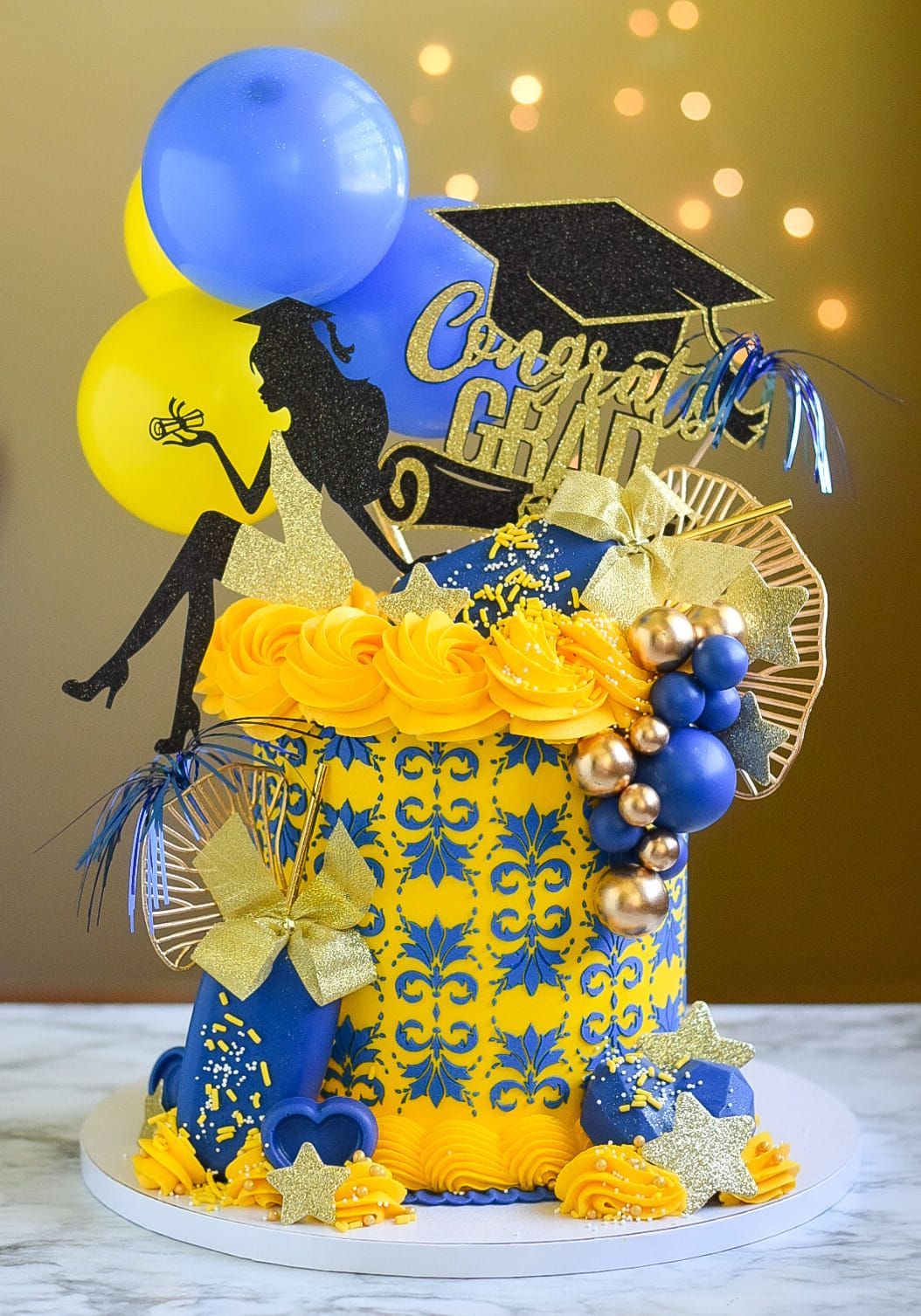 Graduation cake with yellow, blue, and gold decorations, balloons, and a silhouette.