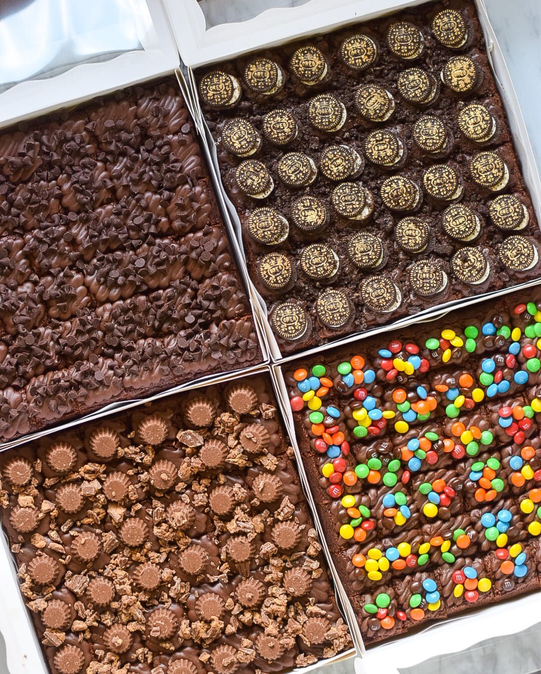Brownie assortment with various toppings, including cookies, candies, and chocolate chips in a white box.