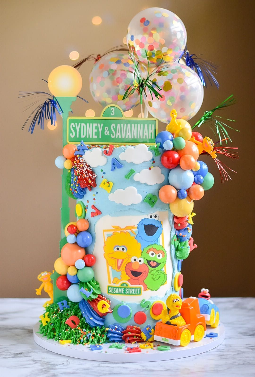 Sesame Street-themed cake with characters and balloon decorations; light blue, orange, and yellow.