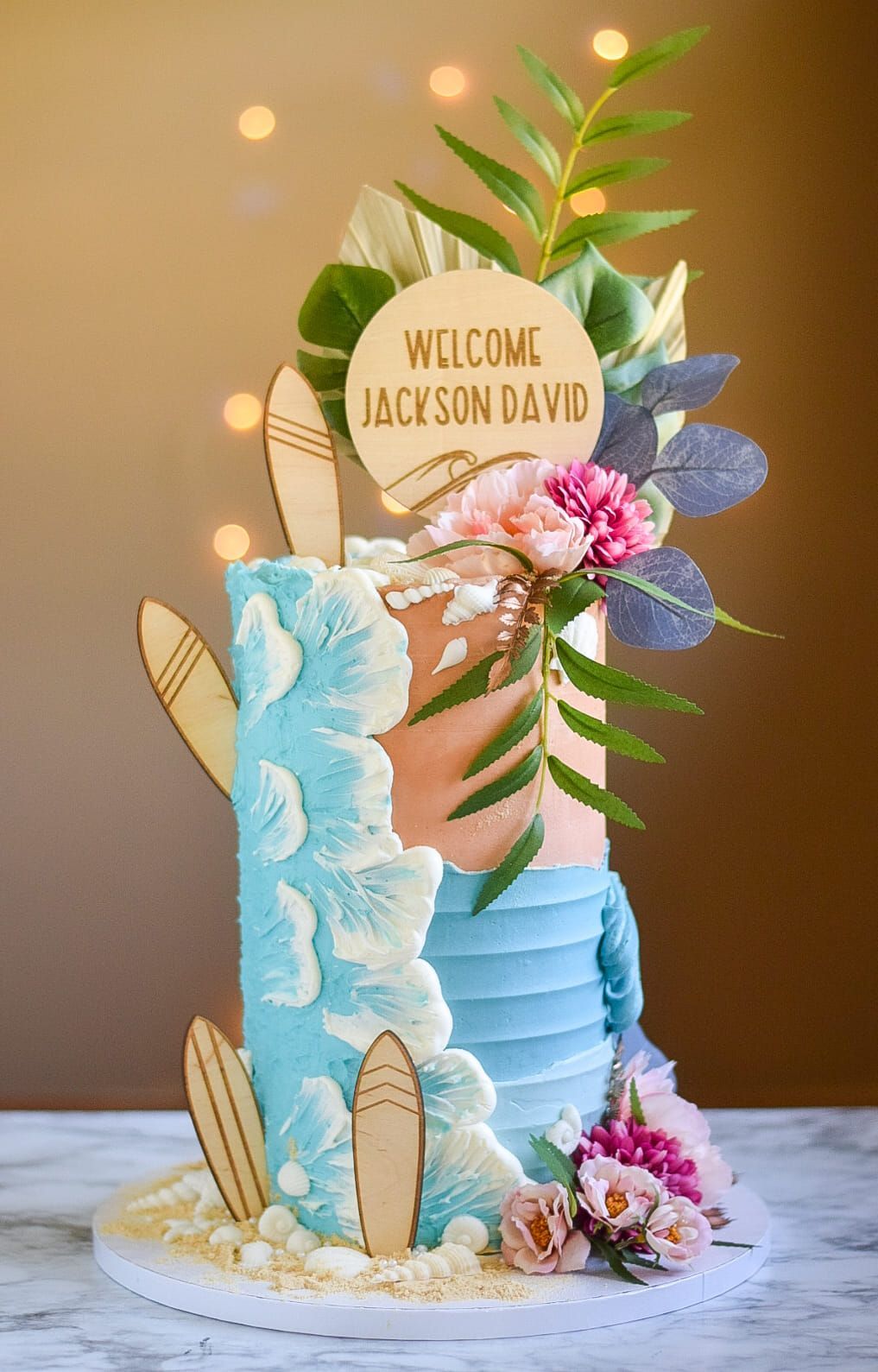 Two-tiered beach-themed cake with pastel frosting, floral accents, wooden surfboards, and a 