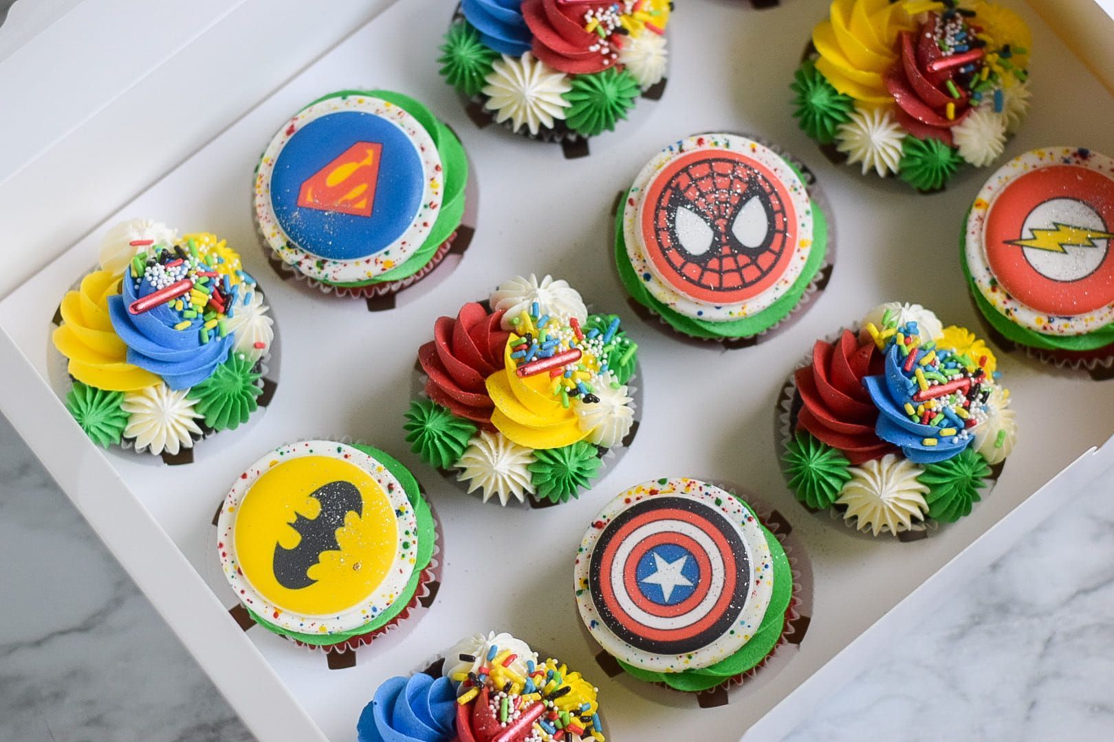 Cupcakes in a box decorated with superhero logos and colorful frosting.