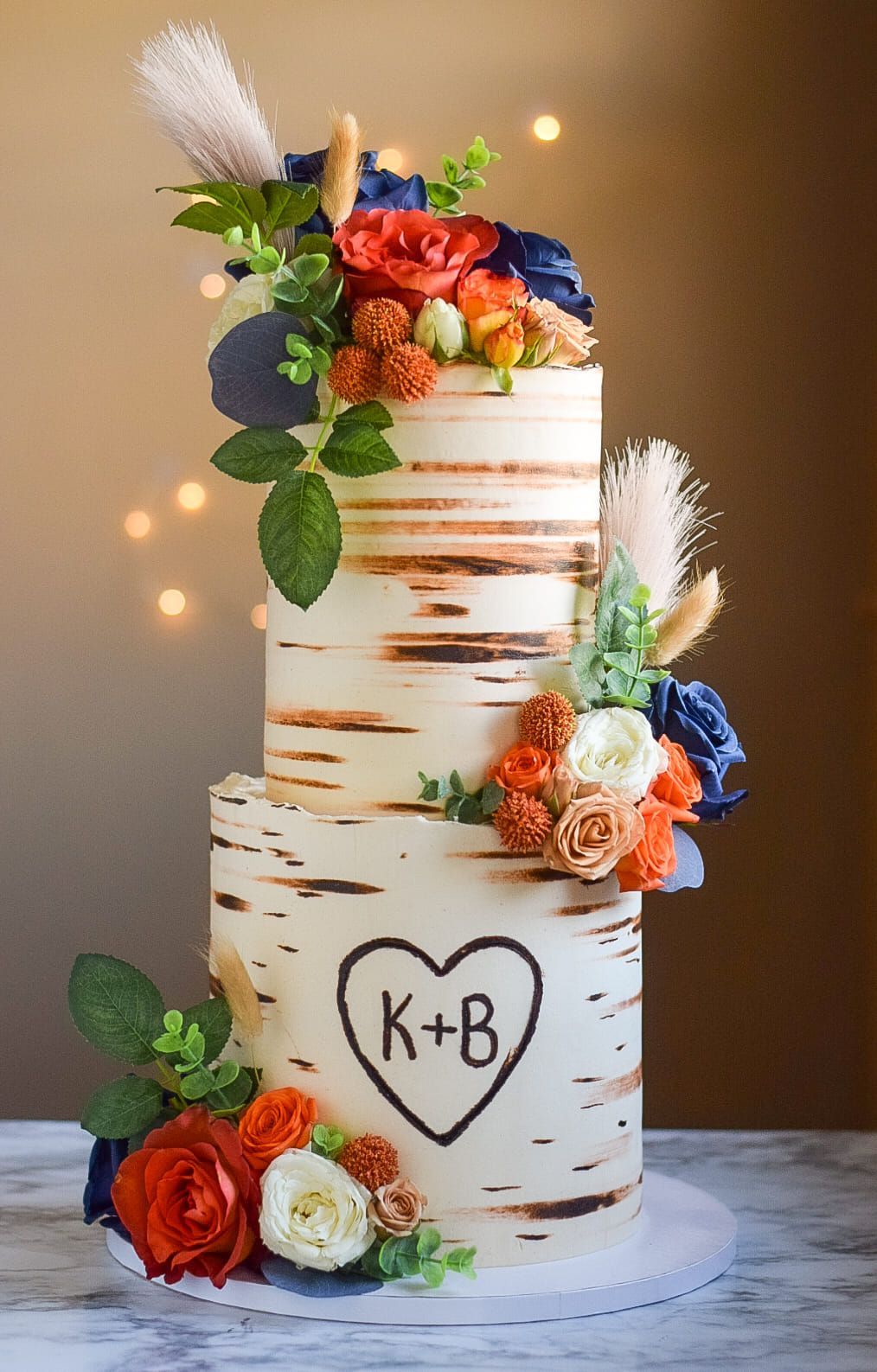 Three-tiered birch-themed cake with floral accents and initials inside a heart.
