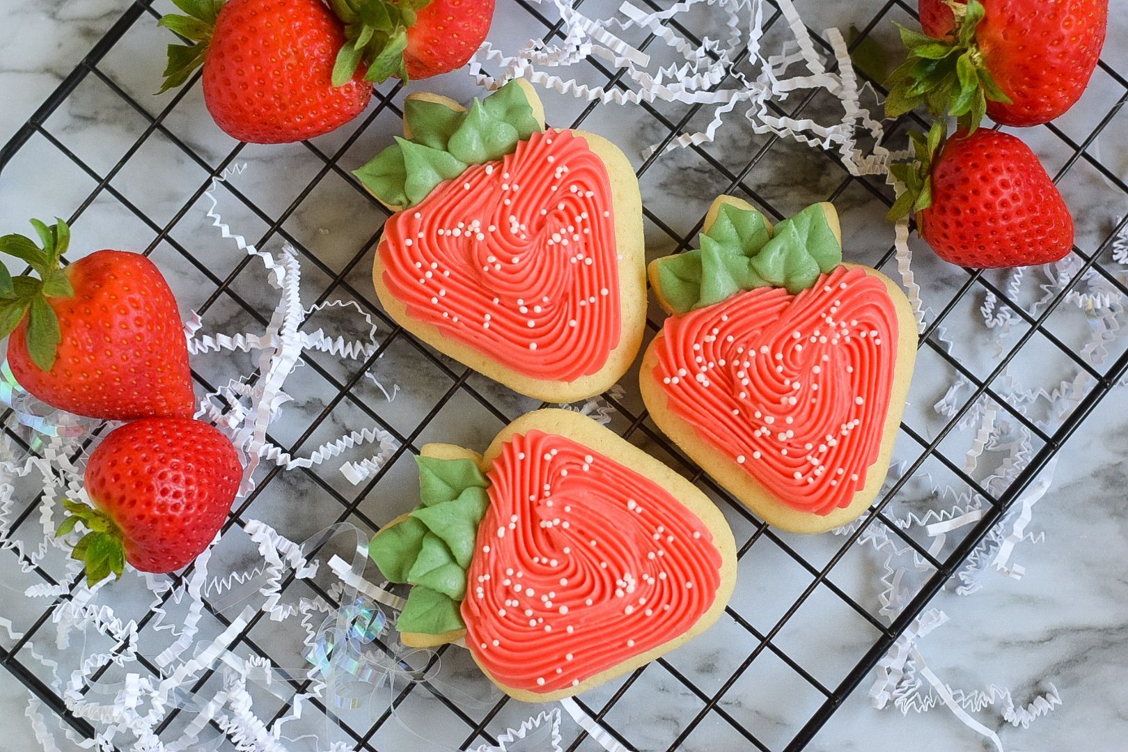 Heart-shaped strawberry pastries on a cooling rack, surrounded by fresh strawberries and white flowers.
