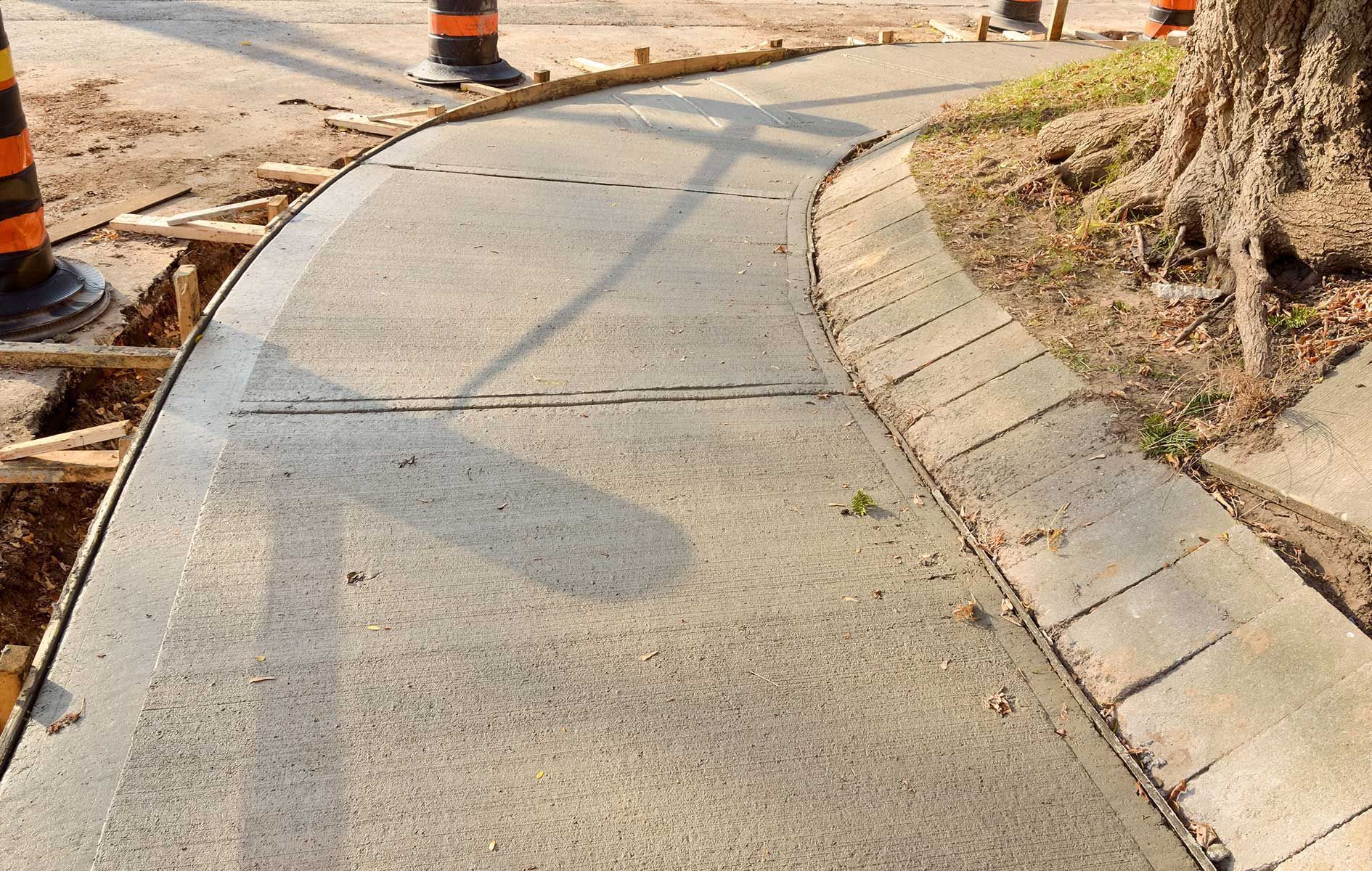 Curved concrete sidewalk beside a dirt embankment and construction cones, with a brick-edged curb.