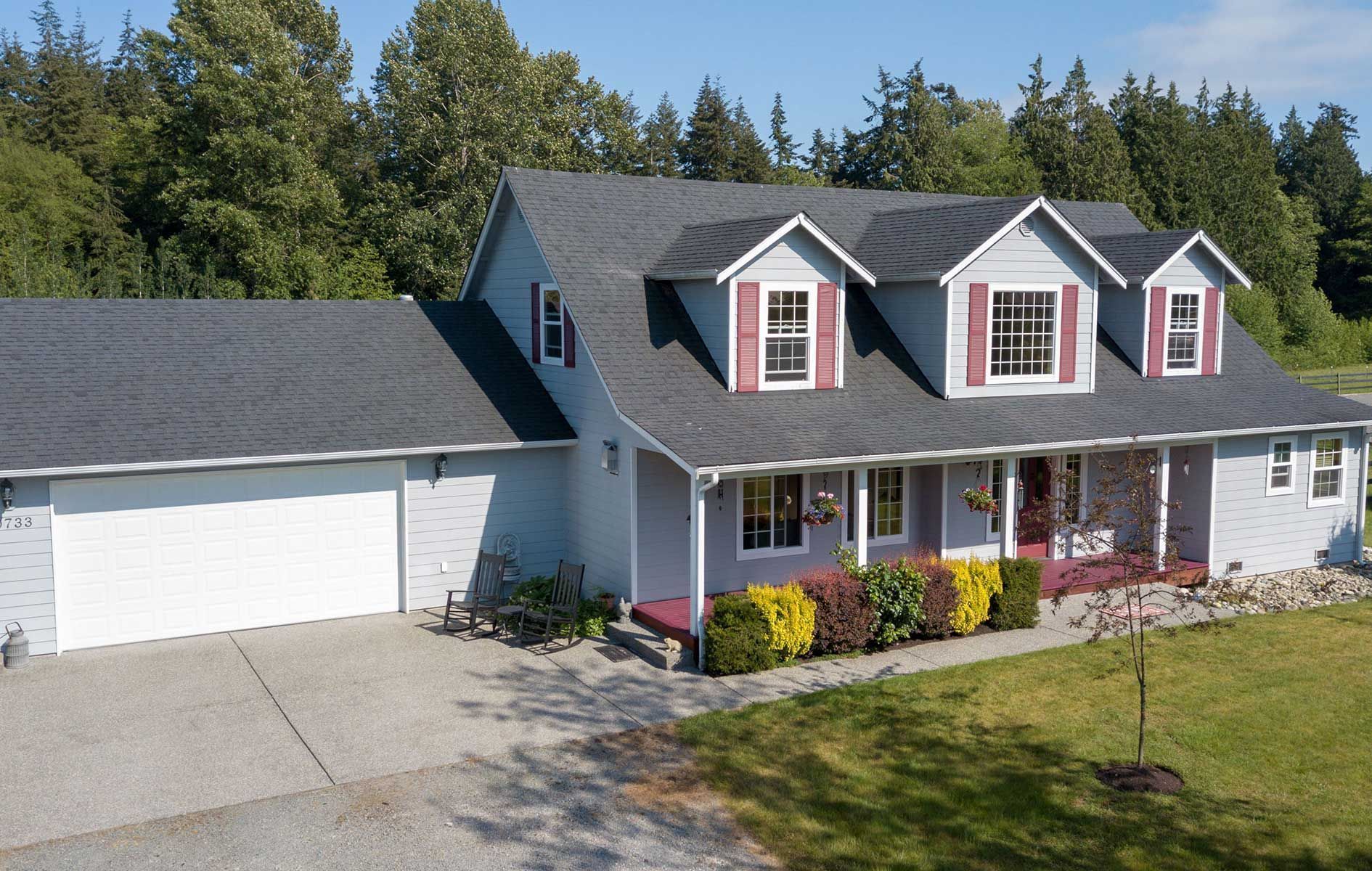 White house with gray roof, red shutters, front porch, and attached two-car garage amid trees and lawn