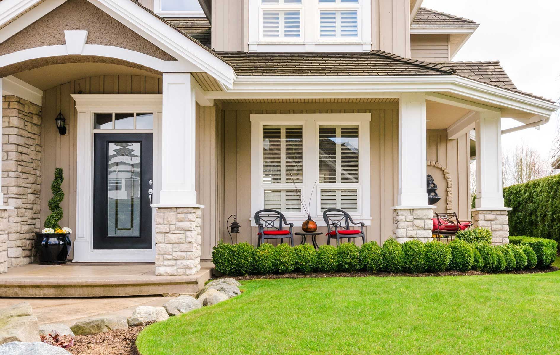 Front porch of a beige suburban house with white columns, stone accents, and red chairs by a green lawn
