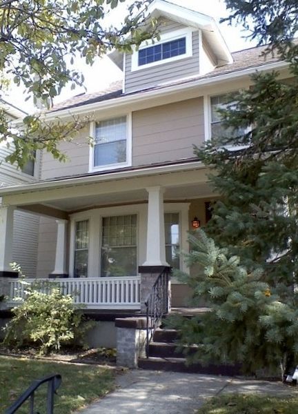 Two-story beige house with a front porch, white columns, and evergreen trees in the yard