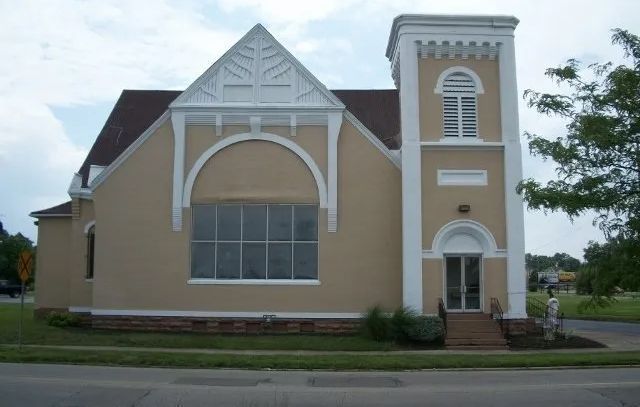 Beige brick building with white trim, arched windows, and a small tower beside a street.