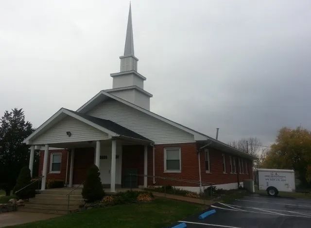 Brick church with white steeple and columns on a rainy day, with a sign by the road