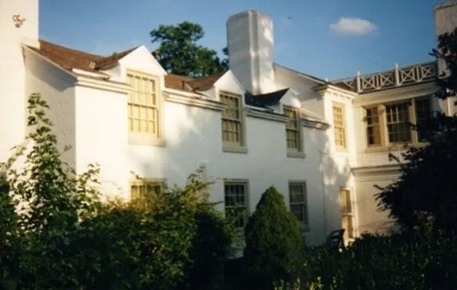 White house with ivy-covered walls, tall windows, and a silver chimney in a sunny garden.