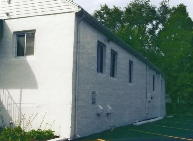 White building exterior beside a parking lot, with windows and trees in the background.