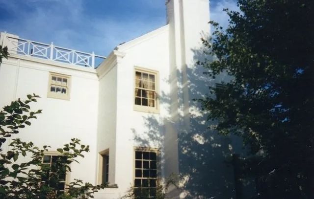 White building facade with windows, rooftop railing, and trees casting shadows in bright sunlight