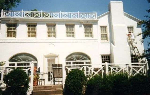 White two-story house with arched windows, balcony railing, and a front garden under bright sunlight