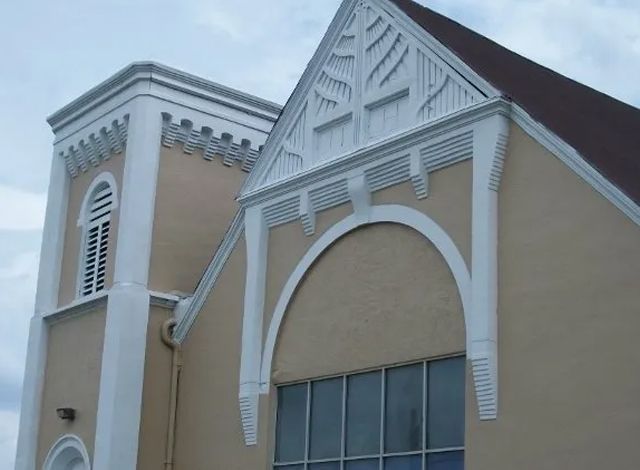 Beige building facade with white trim, arched window, and triangular gable under a cloudy sky