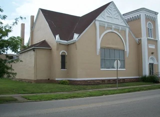 Tan church building with steep roof, arched windows, and beige tower beside a road