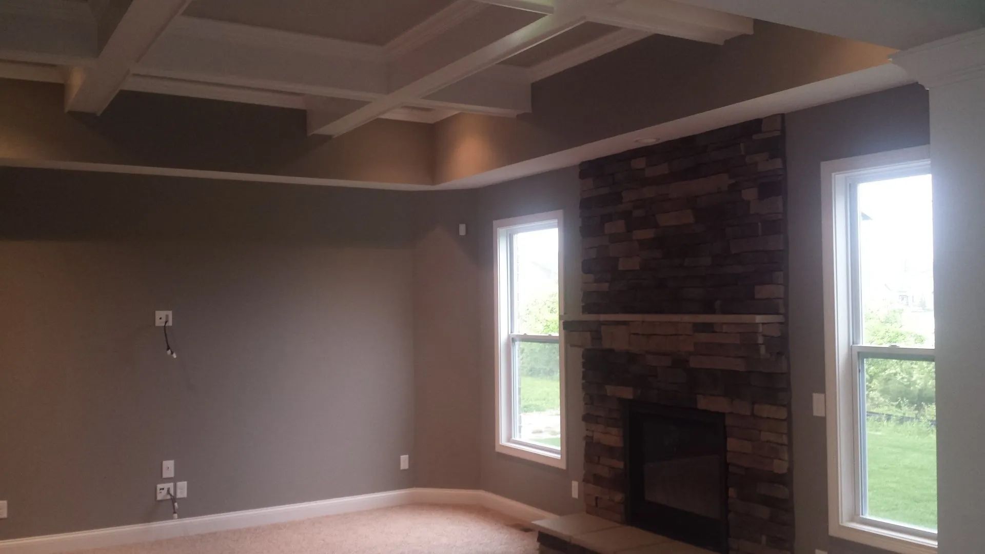 Empty living room with gray walls, tray ceiling, carpet, and a stone fireplace beside two windows.