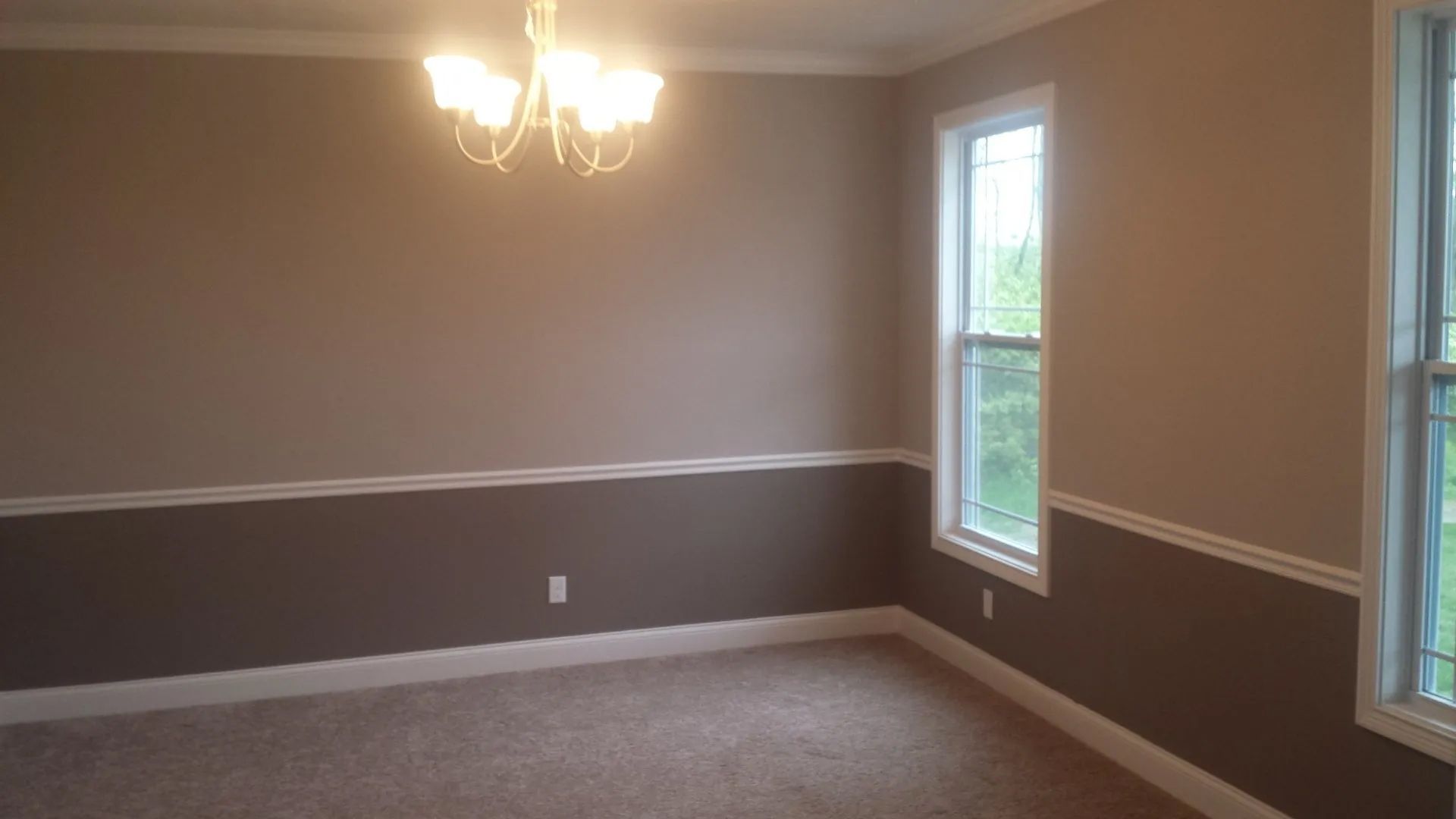 Empty beige dining room with white wainscoting, chandelier, carpet, and two windows.