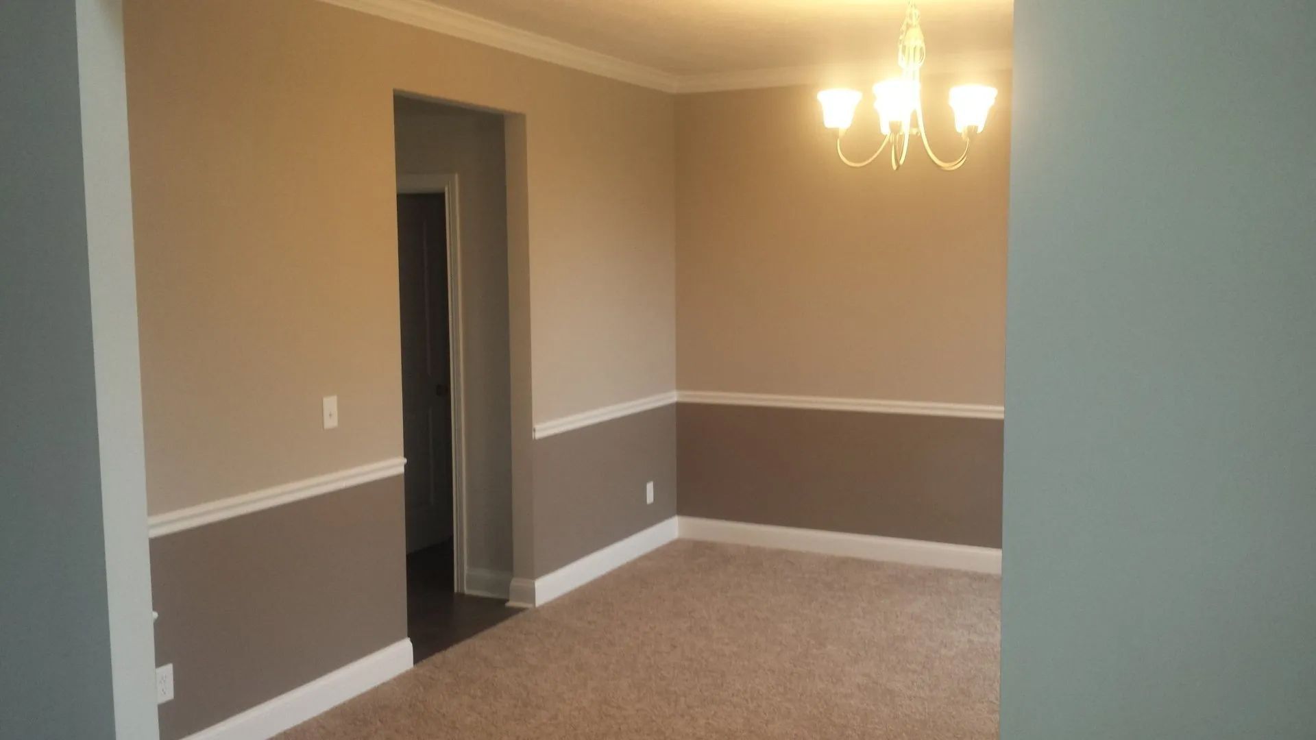 Empty beige dining room with white chair rail, brown trim, carpet, and chandelier lighting
