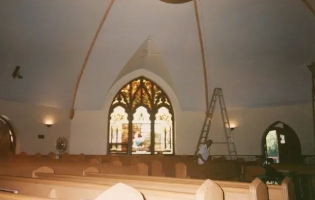Church interior with wooden pews, stained-glass window, and a ladder near the altar area