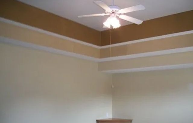 Corner of a beige room with white trim and a ceiling fan under a brown ceiling