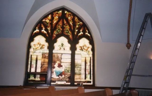 Gothic-style stained glass window in a church, with a ladder leaning against the wall nearby.