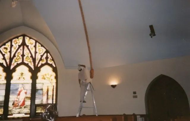 Person on a ladder painting a church ceiling beside an ornate stained-glass window.