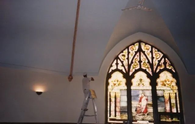 Stained-glass window with ornate arch in a white room, with a ladder and warm wall sconce nearby