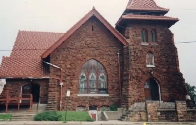 Brick church building with arched stained-glass window and red roof under a cloudy sky