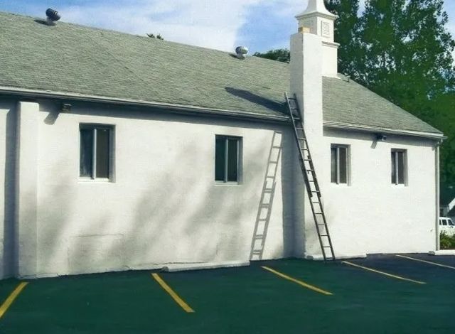 White building with a green roof and two ladders leaning against the wall in a parking lot.