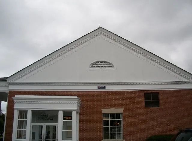 Front of a brick building with a white triangular pediment and cloudy sky above.