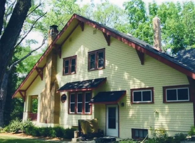 Light yellow house with dark roof and red trim surrounded by green trees