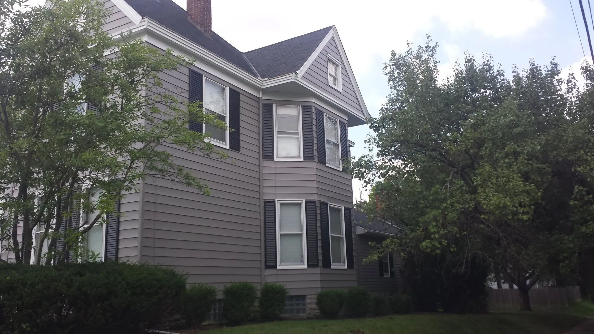 Two-story gray house with white trim and bay windows, partly shaded by trees.