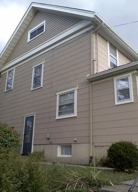 Beige two-story house with gray trim, multiple windows, and a dark front door surrounded by shrubs.