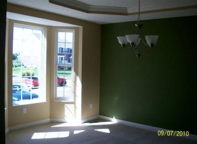 Empty beige room with green accent wall, hanging light fixture, and sunlight streaming through two windows.
