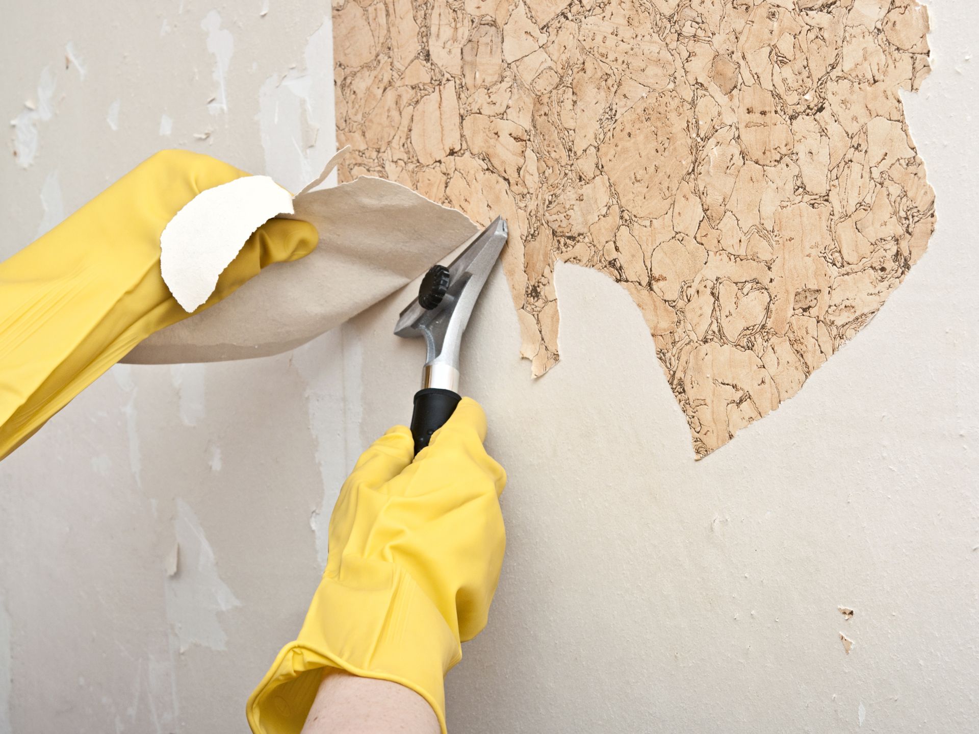 Gloved hands scraping old wallpaper off a wall with a putty knife