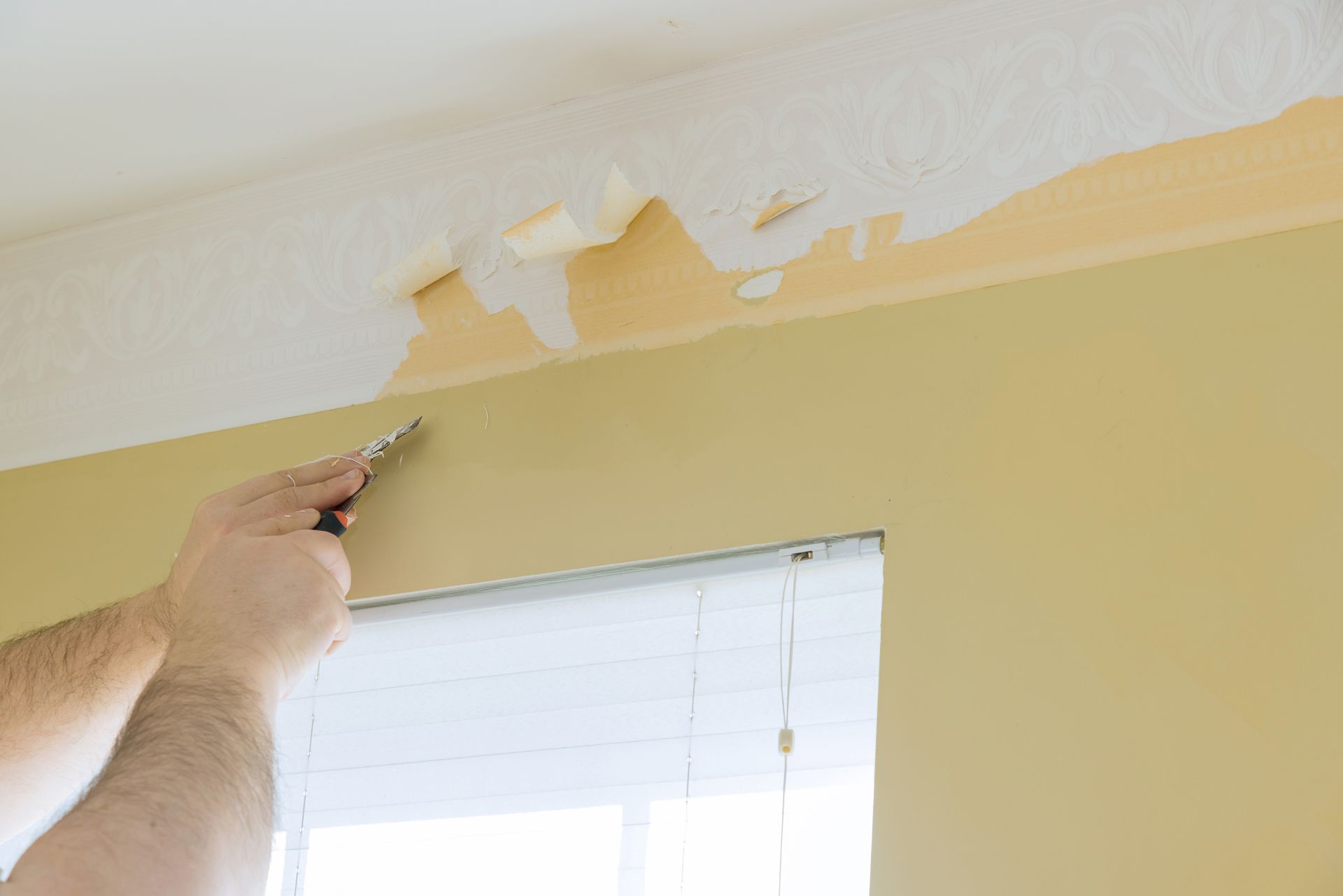 Person scraping peeling paint from a yellow wall above a window with a putty knife.