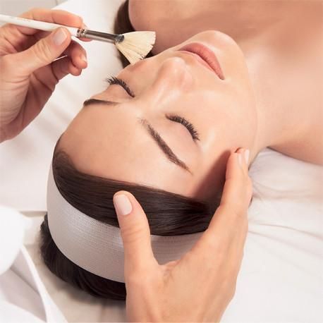 Eine Person, die auf einer Spa-Liege liegt und sich mit einem Pinsel eine Gesichtsmaske auftragen lässt.