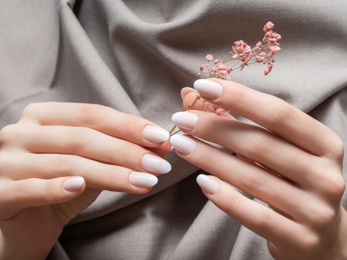 Hände mit weißem Nagellack halten einen kleinen rosa Blütenzweig vor grauem Hintergrund