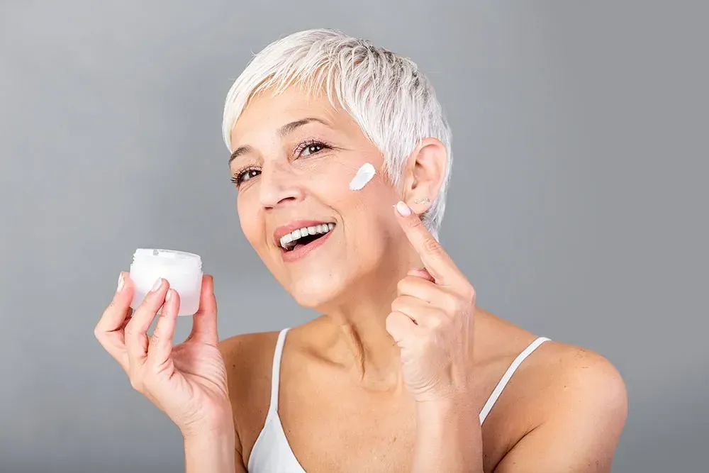 Eine lächelnde Frau trägt Creme auf ihre Wange auf, während sie ein Glas mit Hautpflegeprodukten in der Hand hält.