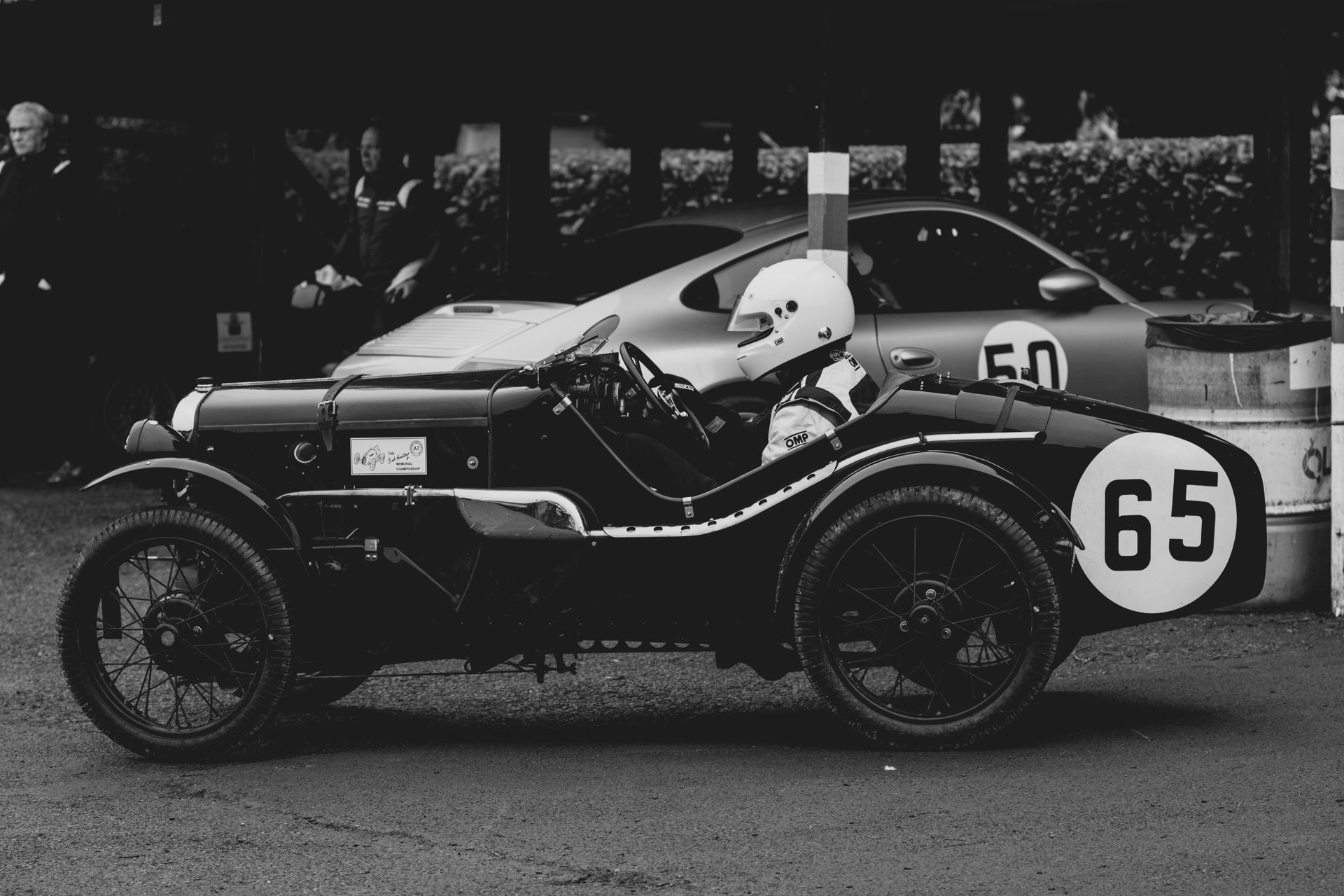 Vintage black race car with number 65 on a street, parked beside another racing car at an event