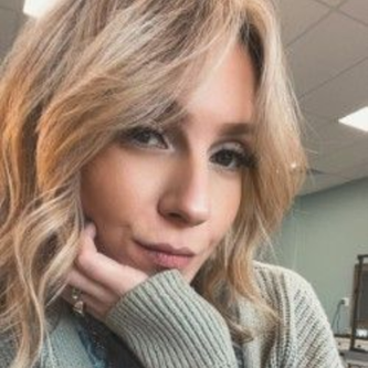 Woman with blonde hair, resting chin on hand, looking at the camera. Wearing a green sweater.