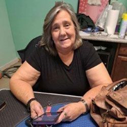 Woman with graying hair and a black shirt, smiling, holding a phone, seated at a table, brown purse beside her.