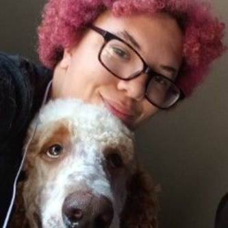 Person with pink hair and glasses smiles with a dog, both looking at the camera.