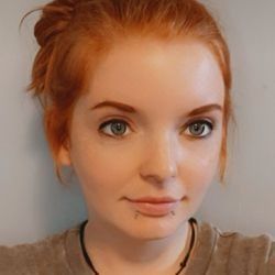 Woman with red hair in a bun, light skin, gray eyes, and lip piercings, smiling slightly.