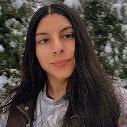 Woman with long dark hair, brown eyes, smiling. Snow-covered evergreens in background.