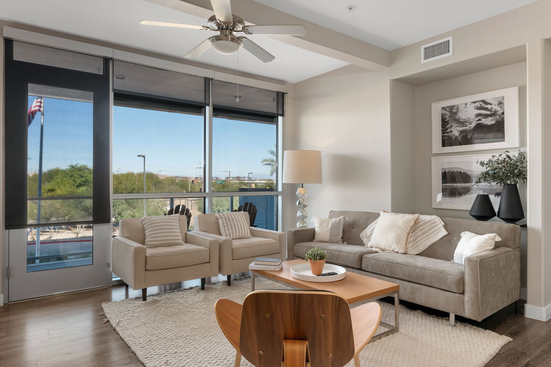 Living room with tan couch, chairs, and wooden coffee table; large windows overlook a city street.