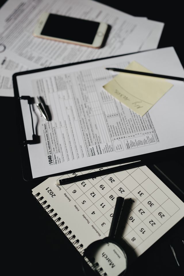 Desk with clipboard paperwork, sticky note, calculator, magnifying glass, and smartphone on a dark table.