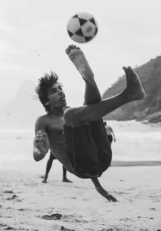 A man is doing a trick with a soccer ball on the beach.